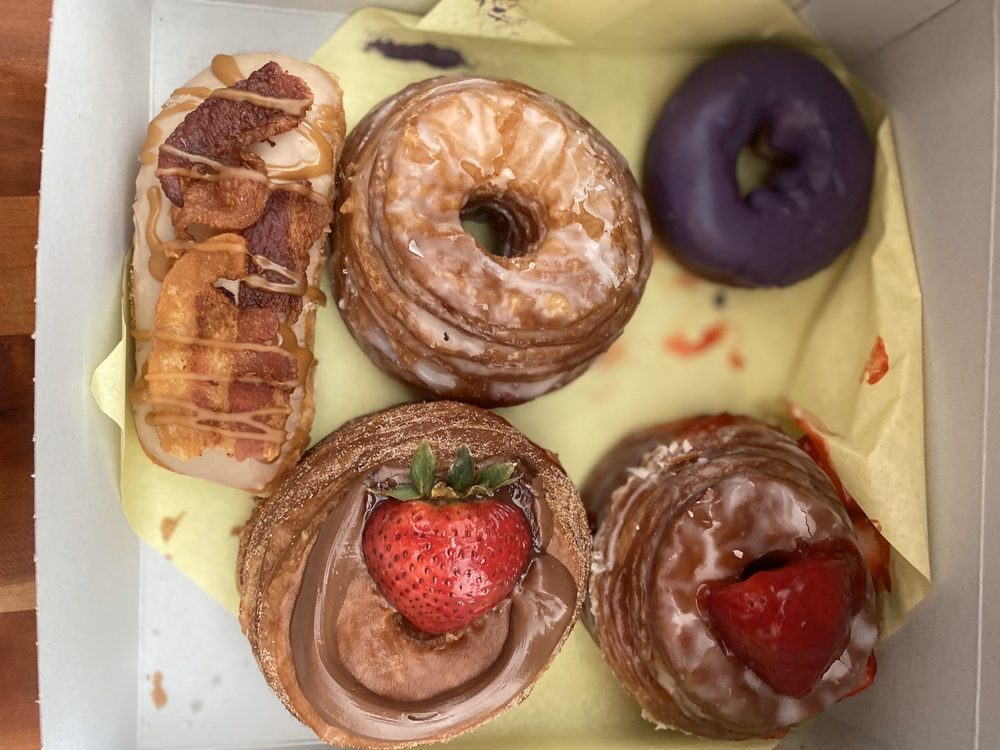 An open box holding five pastries: a bar topped with bacon, a glazed croissant donut, a purple ube donut, a chocolate-filled one with a whole strawberry, and a croissant donut split around strawberries.
