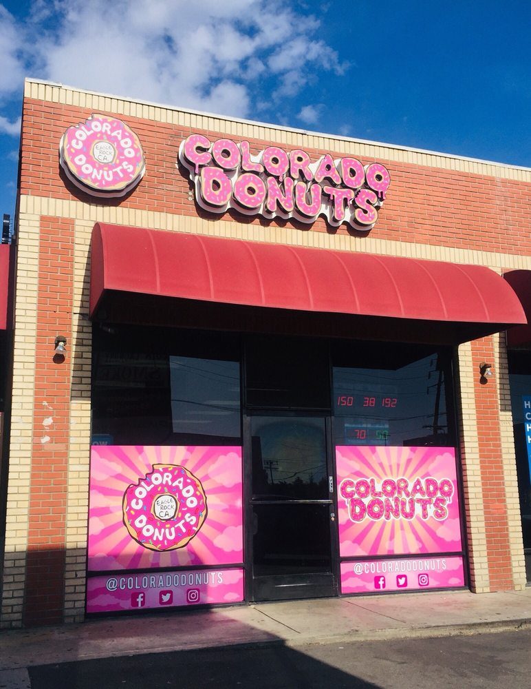 The Colorado Donuts storefront under a blue sky, with a lit pink sign spelled in donut-shaped letters above a red awning and pink starburst window graphics below.