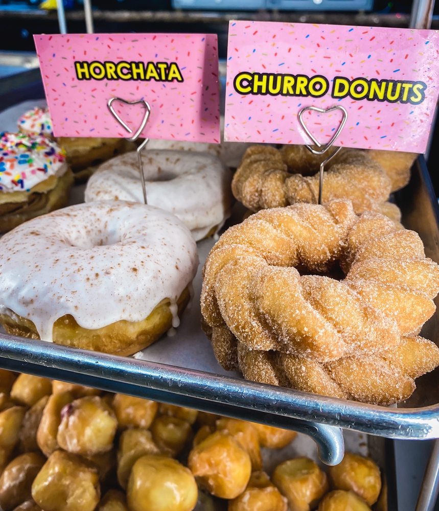 Two pink tags reading horchata and churro donuts standing over a tray of cinnamon-sugar twisted donuts and a white iced donut, with glazed donut holes on the shelf below.