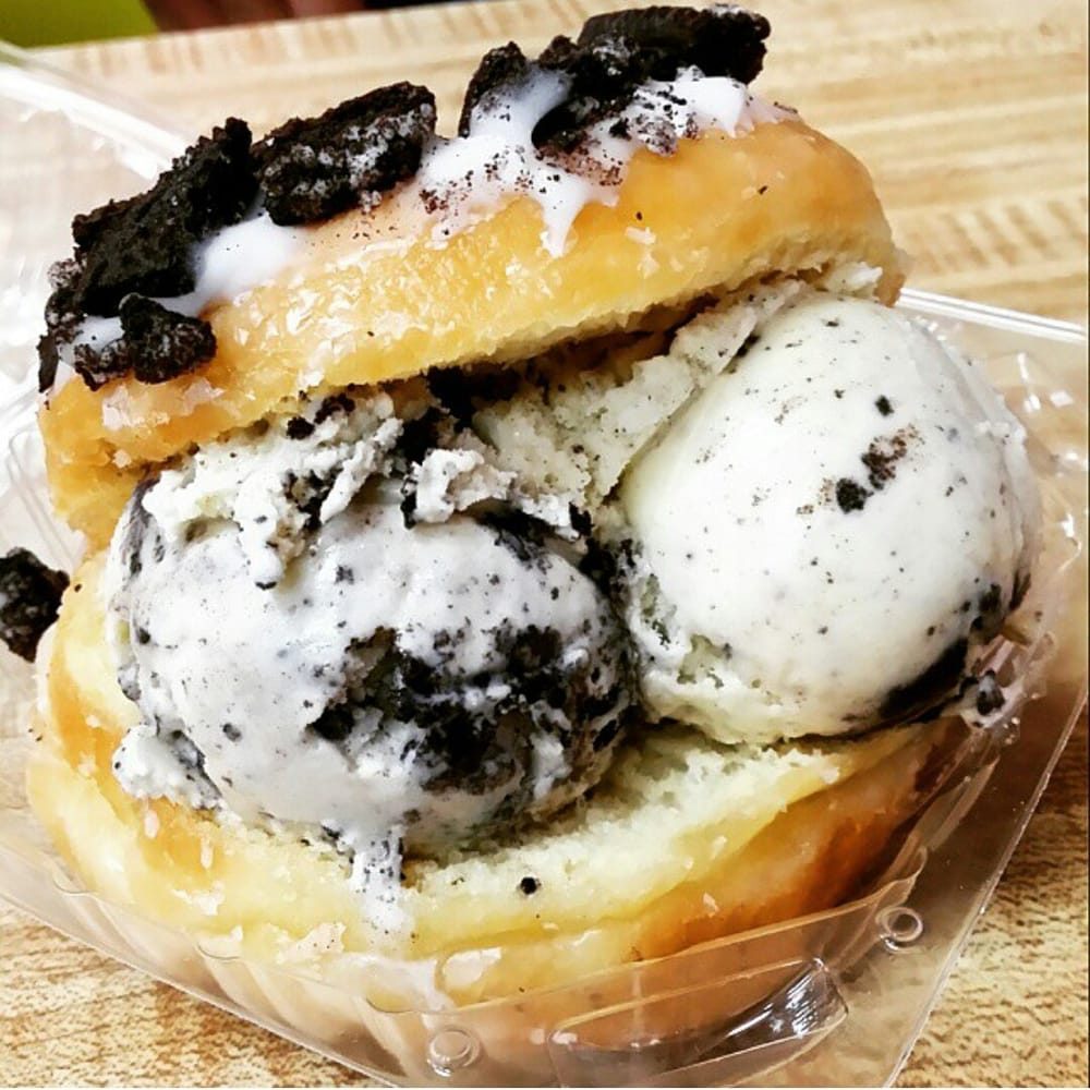 A glazed donut split open around two scoops of cookies-and-cream ice cream, with crumbled chocolate cookie on top, in a clear takeout container.