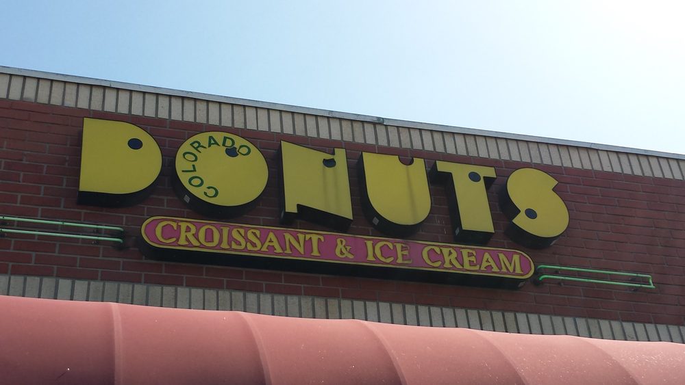 The older roofline sign on the building: tall yellow DONUTS letters with COLORADO written sideways inside the O, above a long rose-pink panel with a yellow border reading croissant and ice cream.