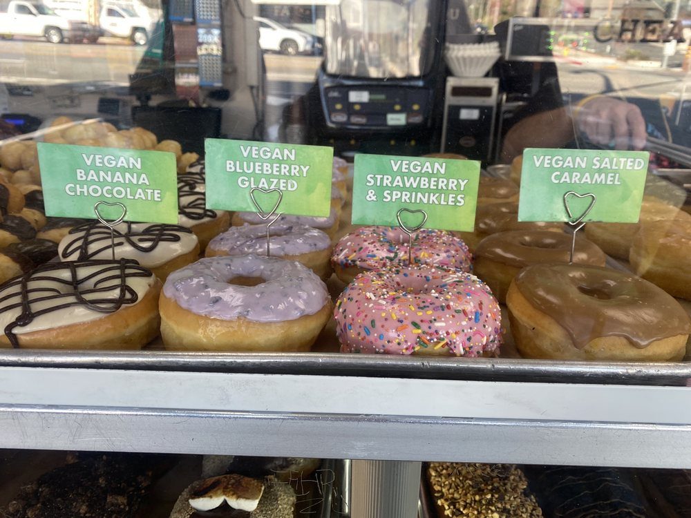 A shelf of donuts behind case glass with green tags reading vegan banana chocolate, vegan blueberry glazed, vegan strawberry and sprinkles, and vegan salted caramel.